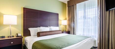 Ein Hotelzimmer mit einem großen Bett, zwei Nachttischen, einem Flachbildfernseher und einem Fenster mit Vorhängen.