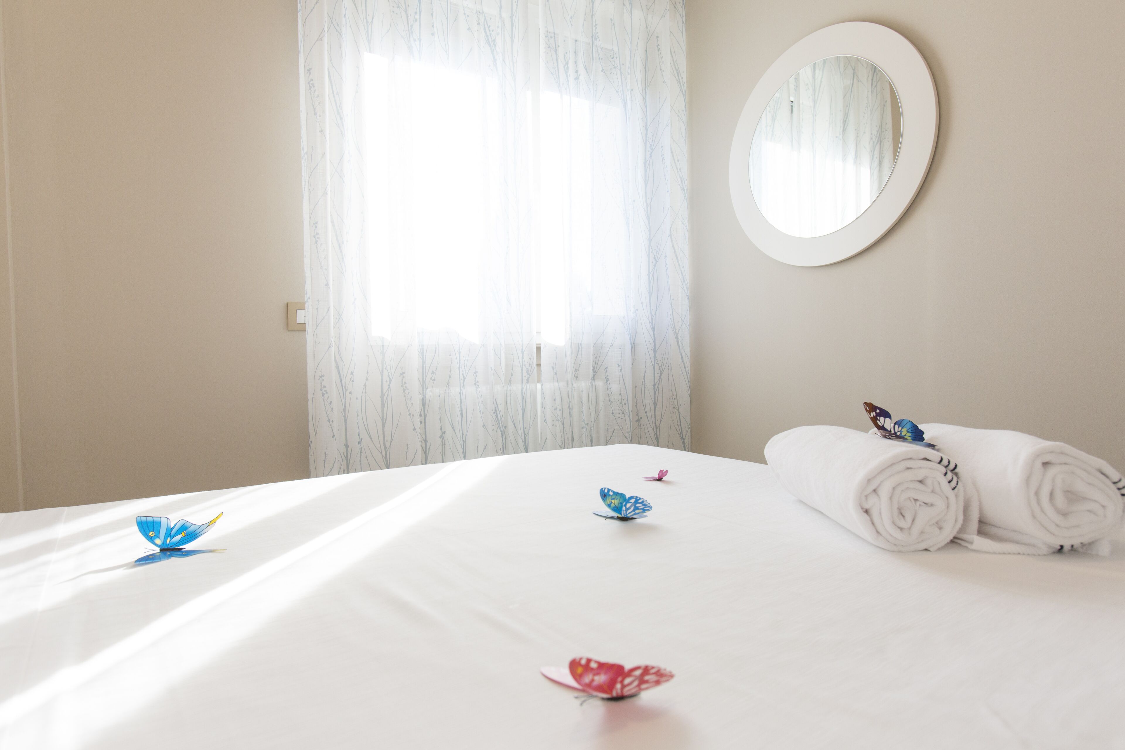 Habitación de hotel con una cama, un espejo redondo y una ventana con cortinas translúcidas.