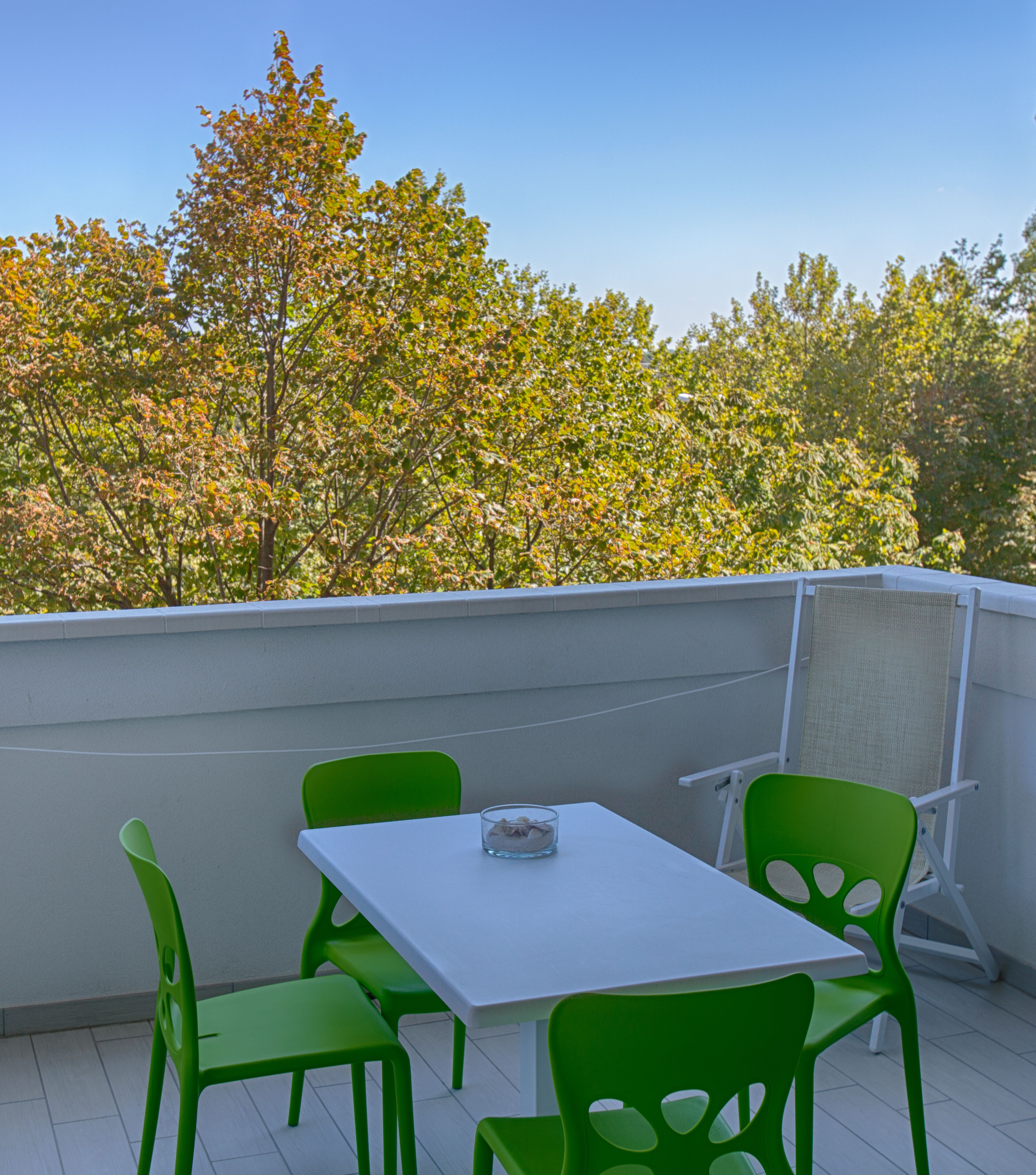 A balcony with a white table and green chairs, overlooking a tree-lined area.