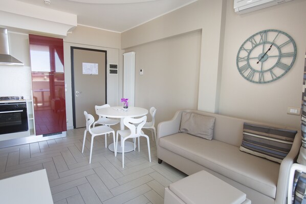 A modern living room with a beige sofa, a round table with chairs, a clock on the wall, and a kitchen area with a red door.