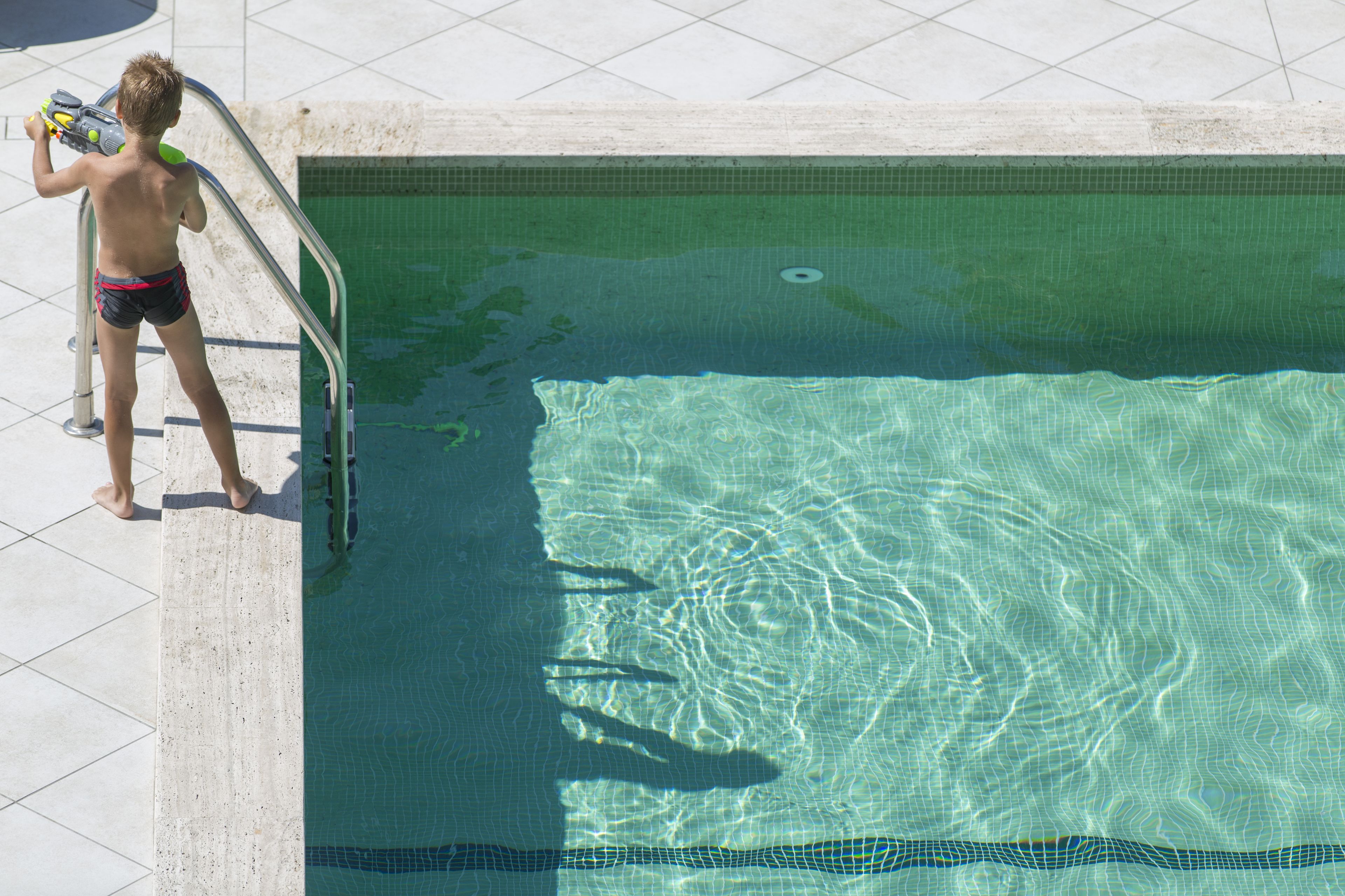 Ein Kind steht am Beckenrand eines Schwimmbeckens mit klarem Wasser und einer gefliesten Terrasse.