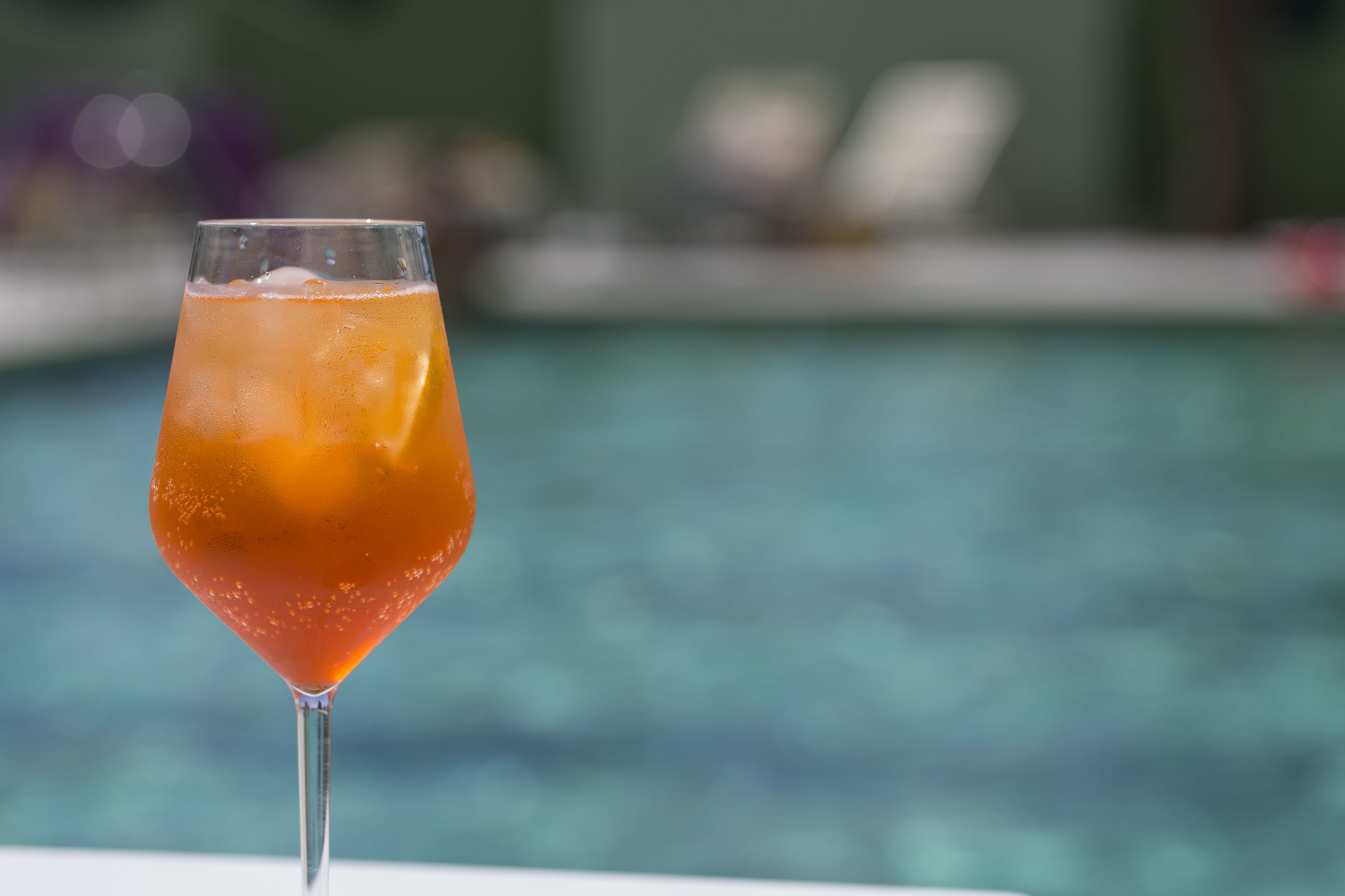 Ett cocktailglas med en klar vätska och isbitar, placerat på en vit yta nära en pool med en suddig bakgrund.