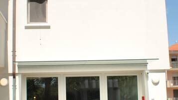 Un bâtiment moderne avec une façade blanche, une entrée en verre et deux plantes en pot placées de part et d’autre de l’entrée.