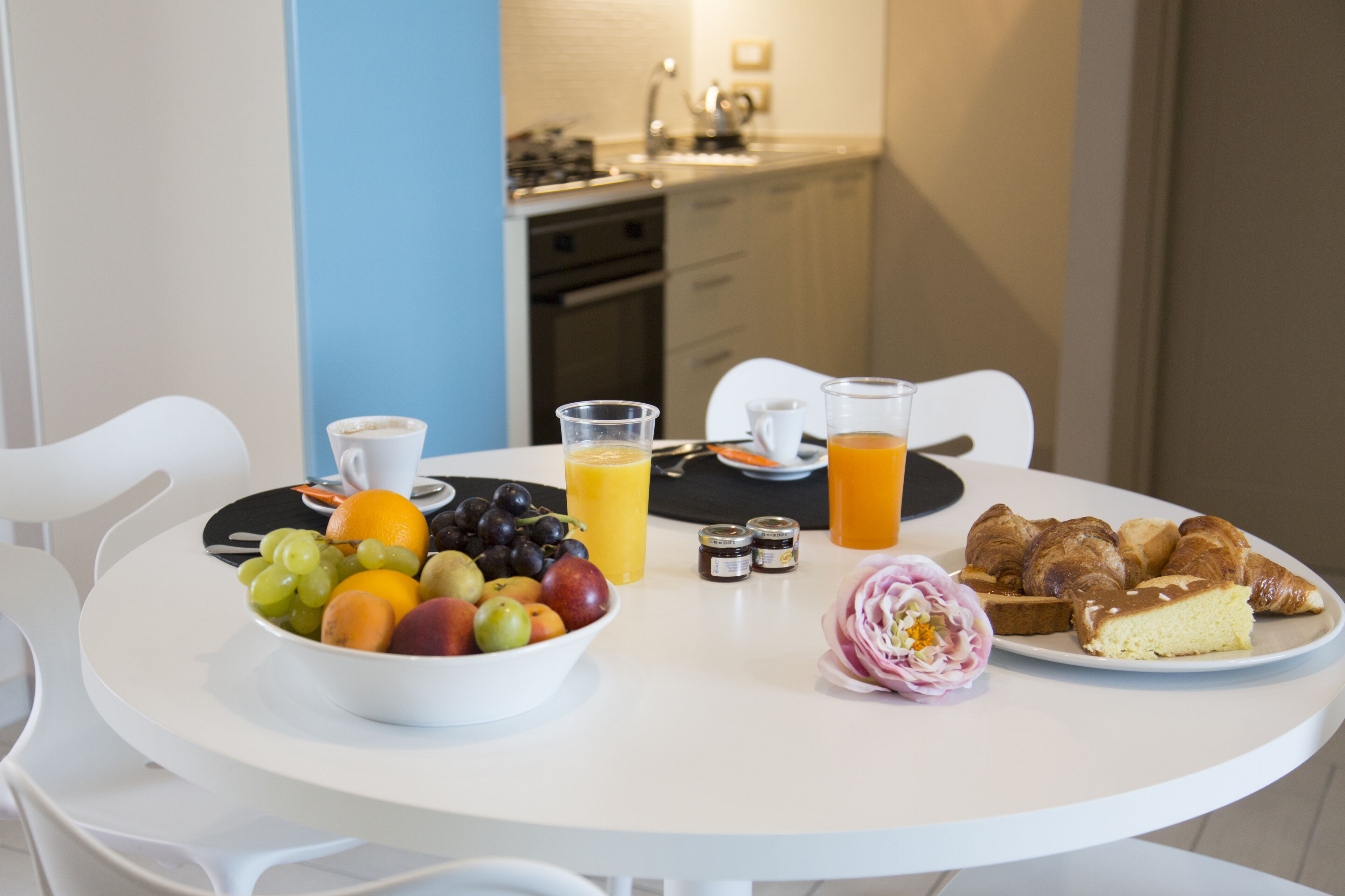A breakfast table with fruits, juice, croissants, and a flower.