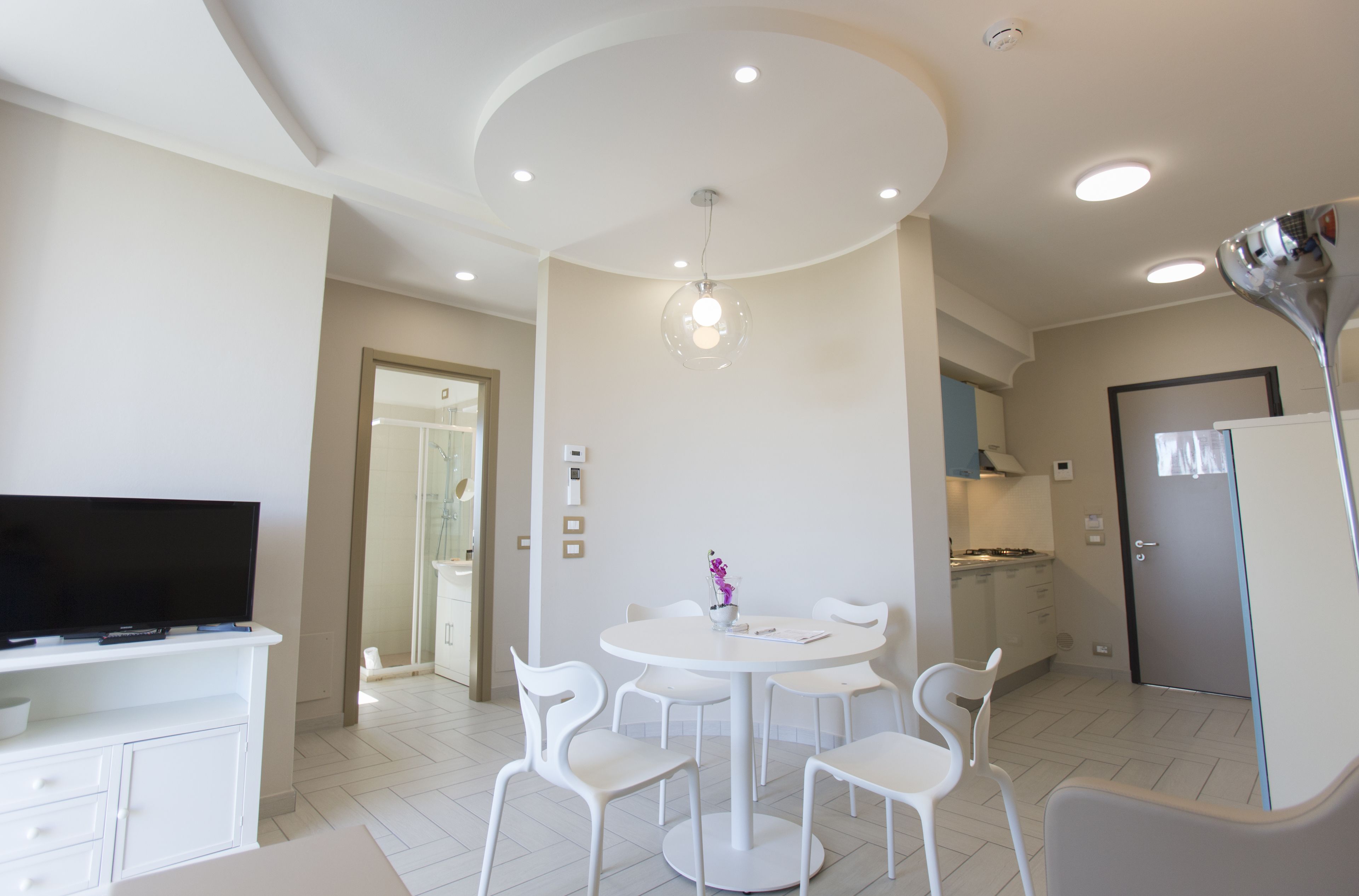 Cozinha moderna com armários brancos, sala de jantar com mesa e cadeiras redondas, e televisão de tela plana sobre um armário branco.