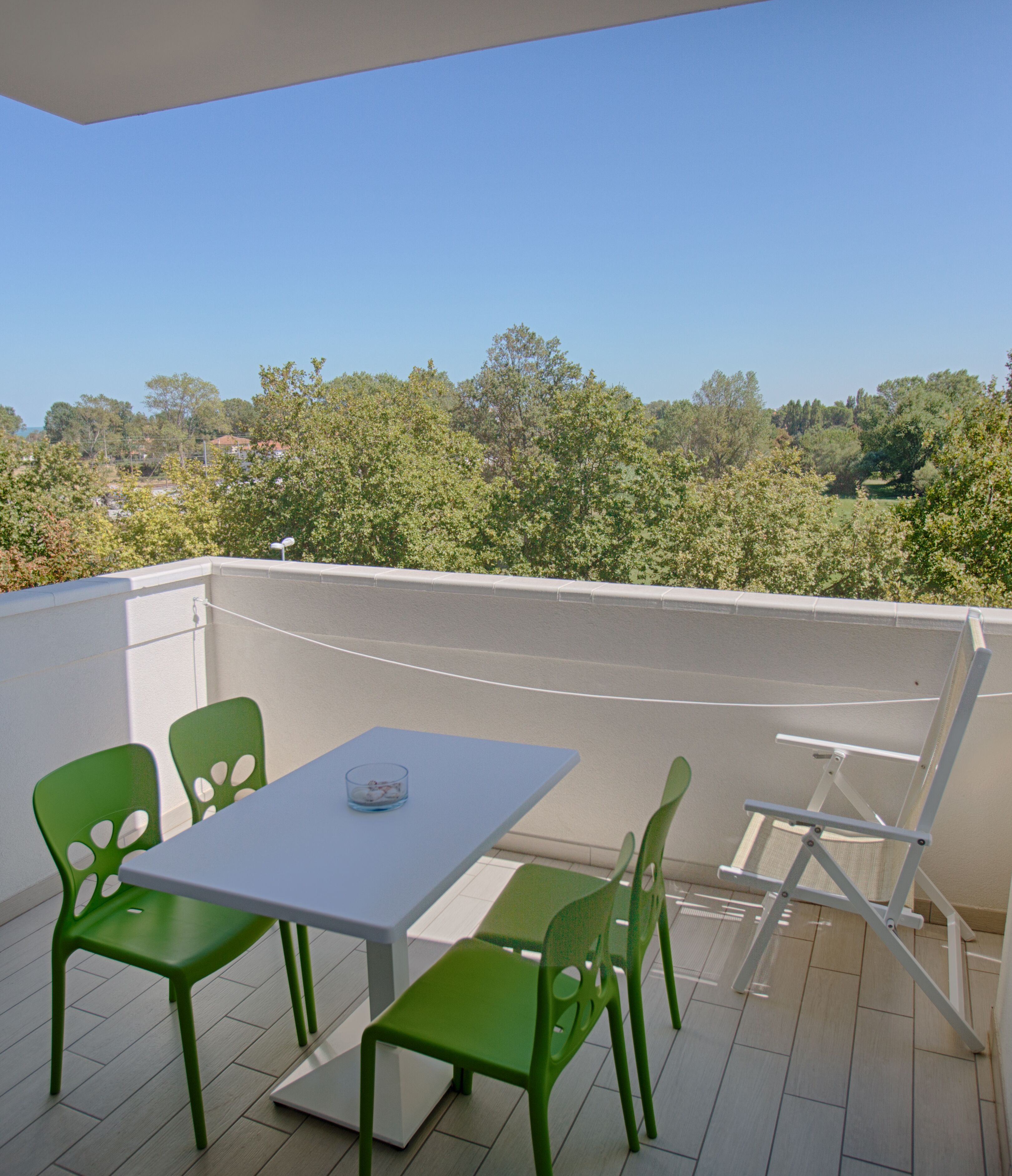 A balcony with a table and four green chairs, overlooking a view of trees and houses.