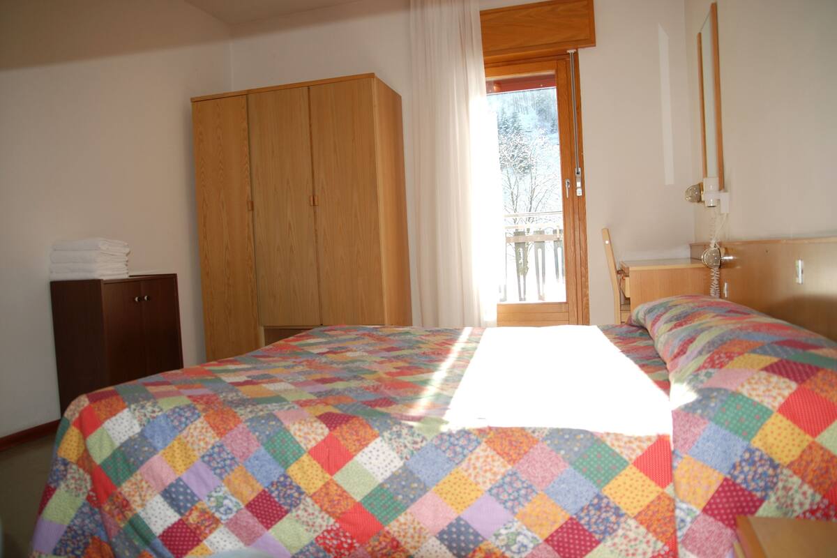 A bedroom with a colorful quilted bedspread, a wooden wardrobe, and a window with curtains.