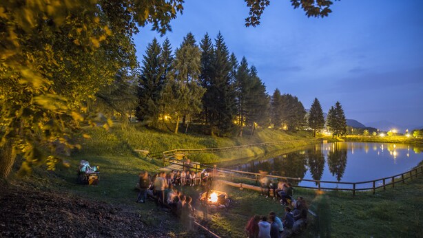 A lakeside gathering with a campfire, surrounded by trees and a wooden fence. - Hotel Bucaneve (Brentonico)