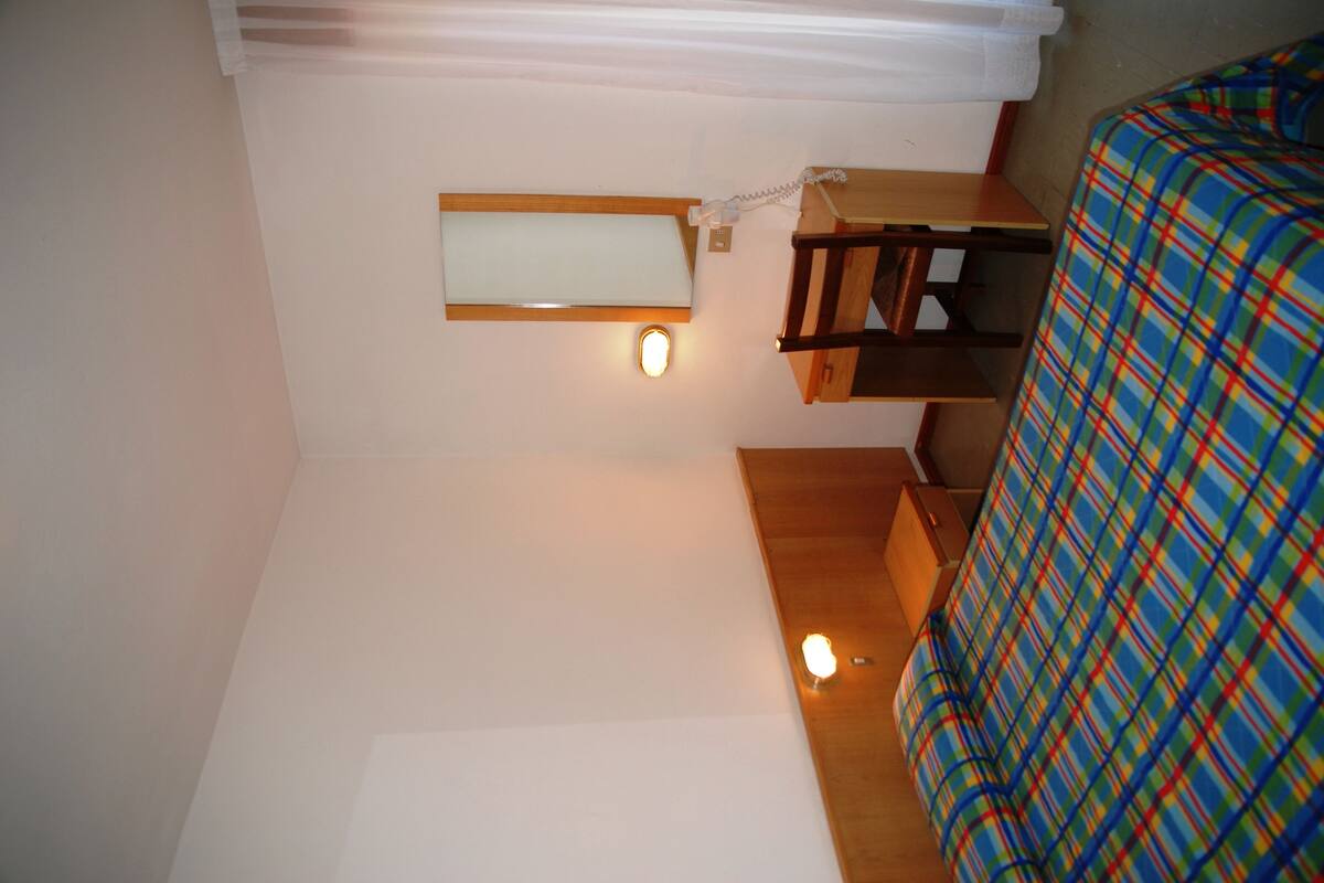 A small, compact room with a bunk bed, a bedside table, a nightstand, and a wall-mounted light fixture.