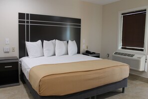 A hotel room with a large bed, a nightstand, a bedside table, a window with blinds, and a wall-mounted air conditioning unit.