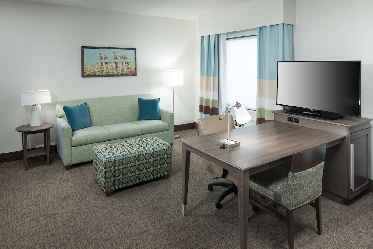 A hotel room with a green sofa, a patterned ottoman, a wooden desk with a television, and a lamp.
