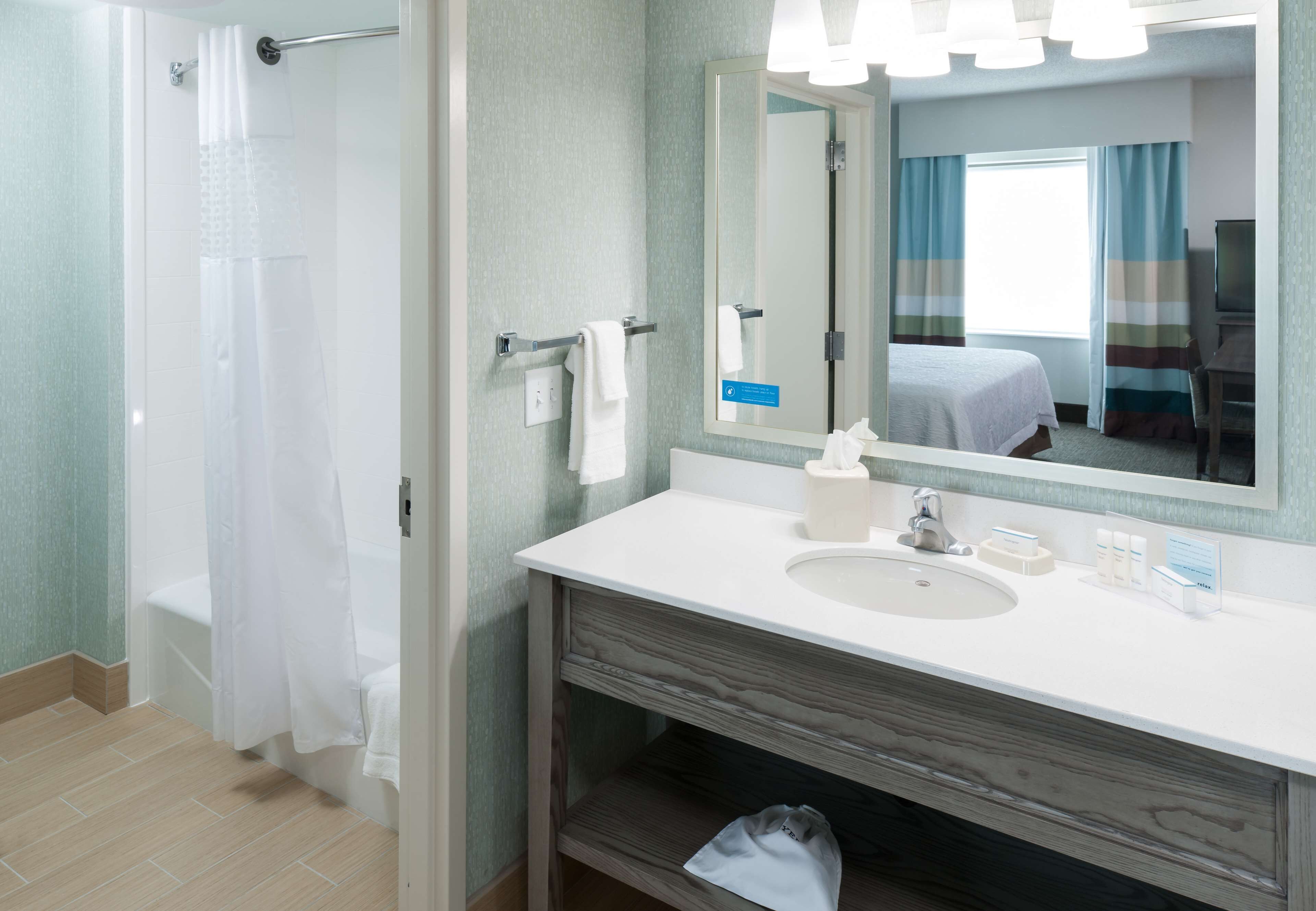 Un baño con ducha, lavabo, espejo y una cama visible en el reflejo.