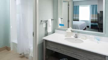 Un baño con ducha, lavabo, espejo y una cama visible en el reflejo.