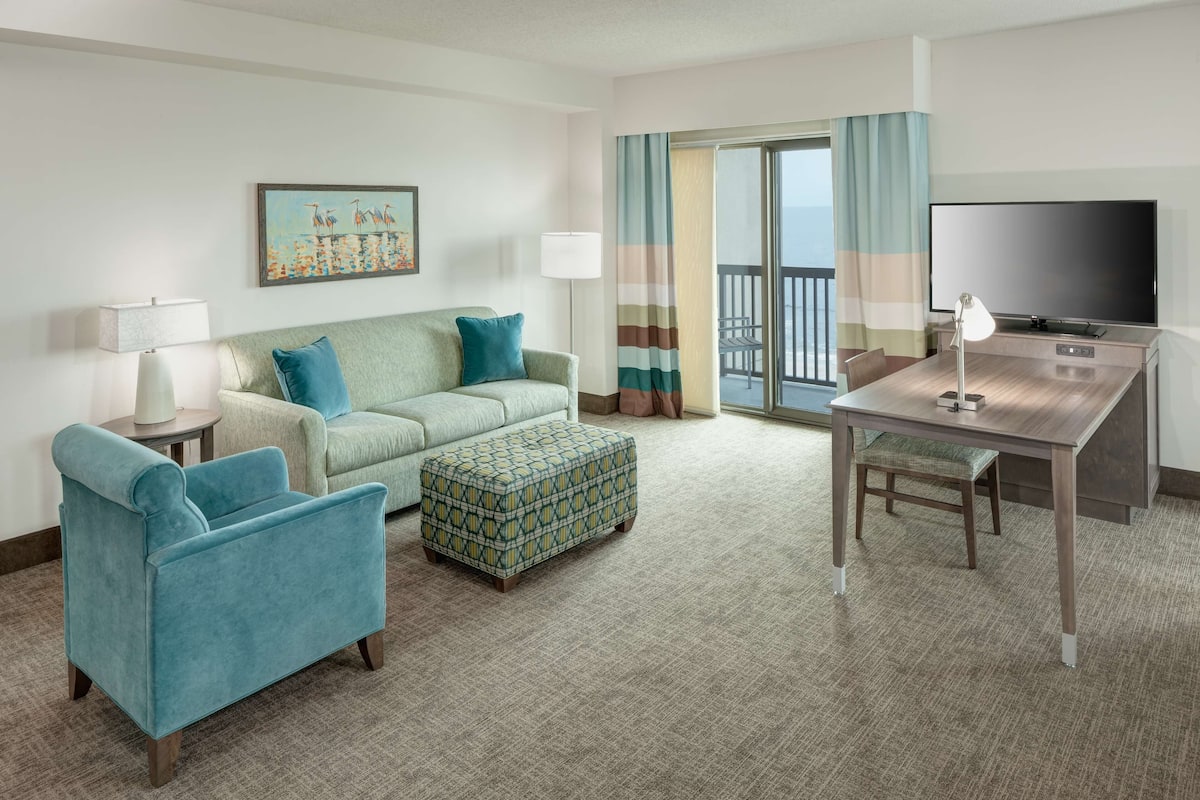 A hotel room with a sofa, armchair, ottoman, desk, TV, and a balcony with curtains.