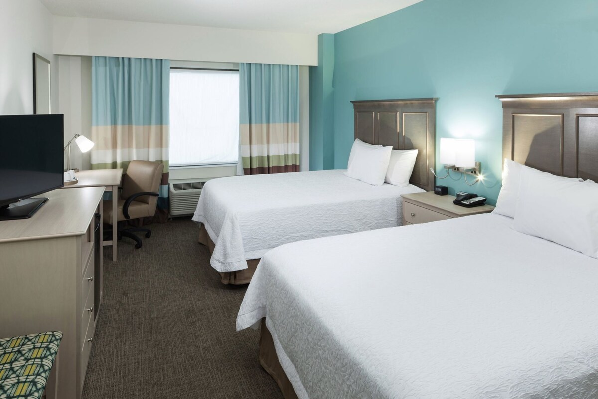 A hotel room with two beds, a desk with a computer, a chair, a TV, and a window with curtains.