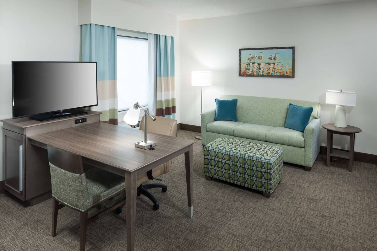 A hotel room with a flat-screen TV, a desk with a lamp, a green sofa with a patterned ottoman, and a painting on the wall.