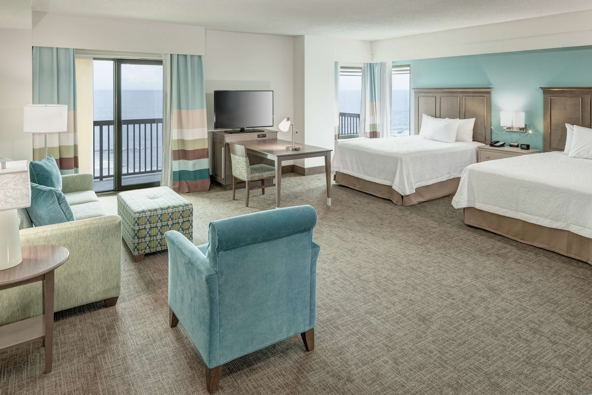 A hotel room with two beds, a desk with a TV, a sofa, and a balcony with curtains.
