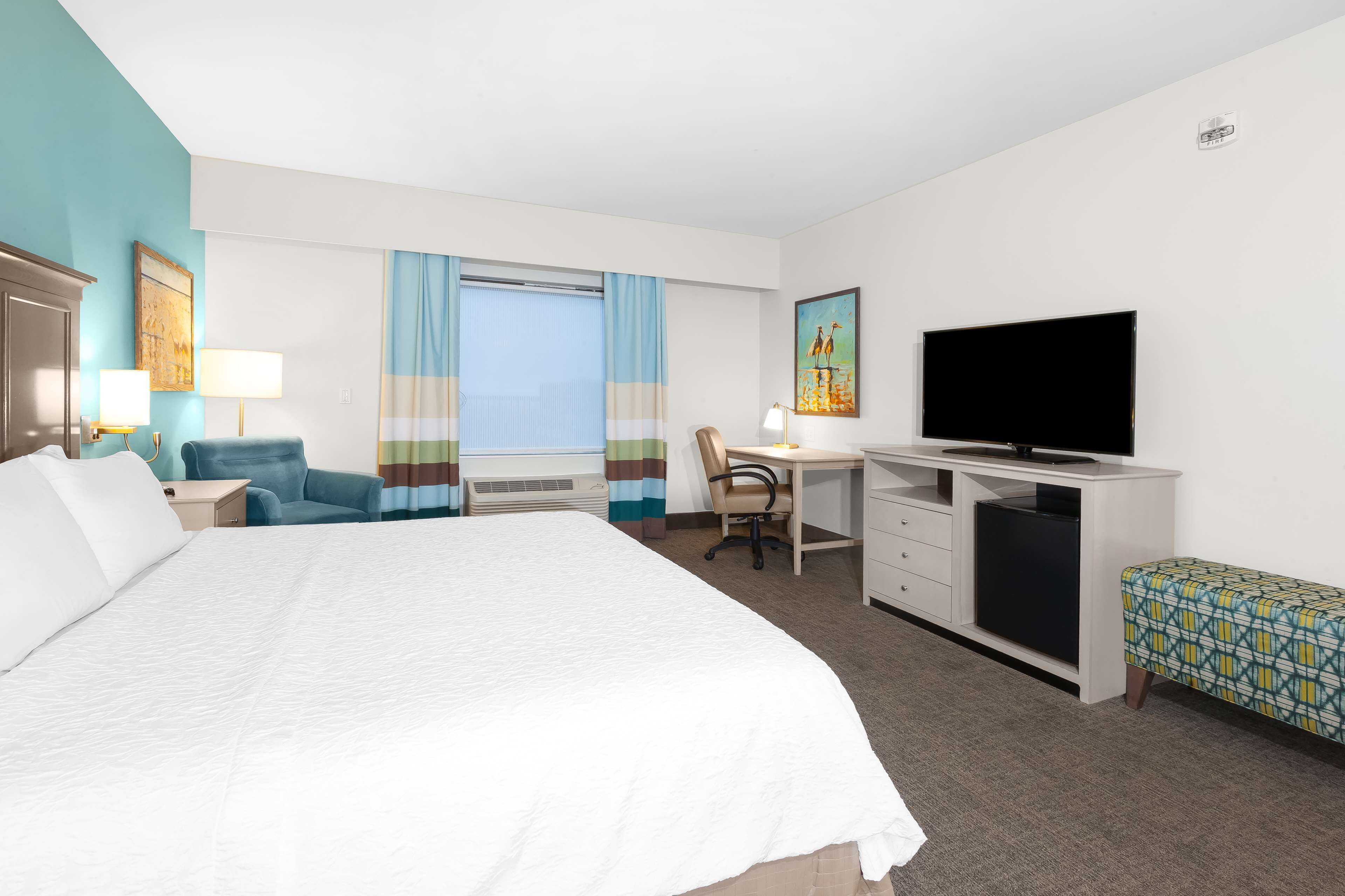 Quarto de hotel com uma cama grande, uma escrivaninha com cadeira, uma televisão e um pequeno banco.