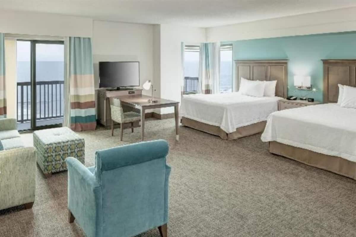 A hotel room with two beds, a desk, a TV, and a balcony with curtains.