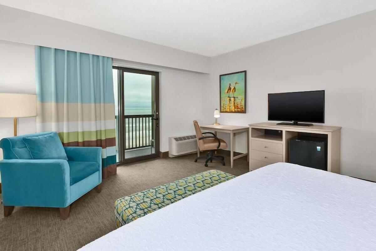 A hotel room with a bed, a desk, a chair, a television, and a view of the ocean through the window.