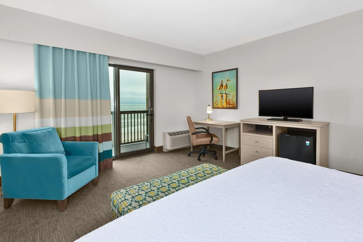 A hotel room with a bed, a desk with a computer, a chair, a TV, and a view of the ocean through a sliding door.
