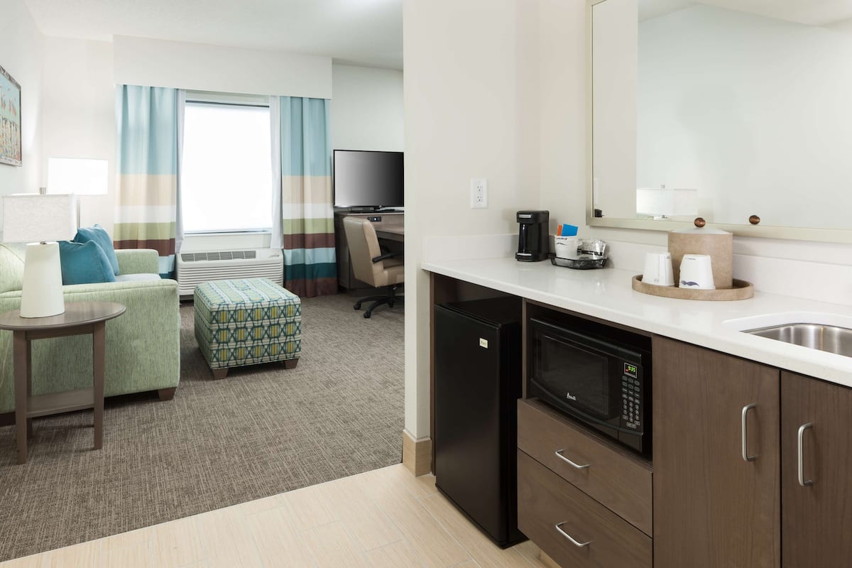 A compact hotel room with a kitchenette, a sofa, a small table, a TV, and a window with curtains.