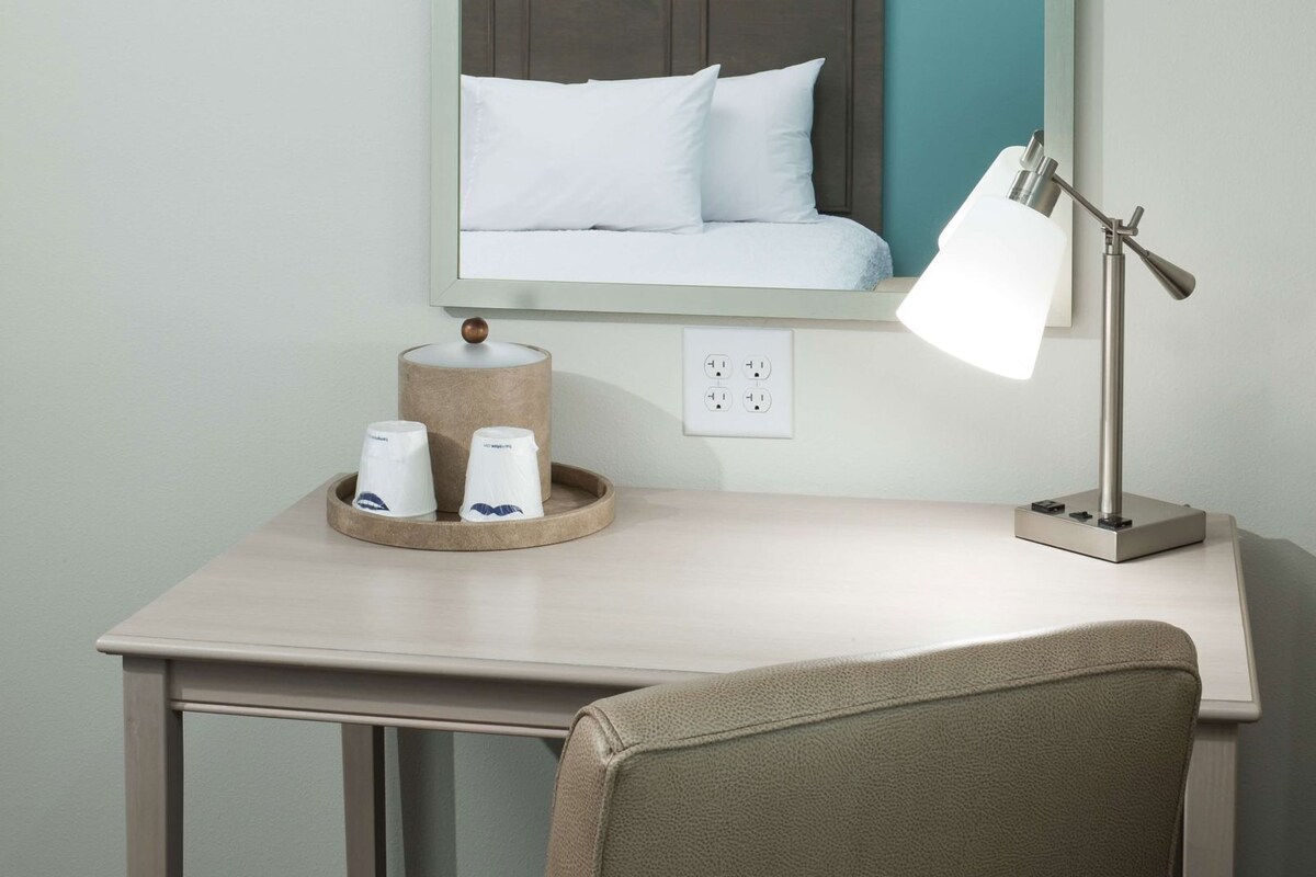 A hotel room desk with a lamp, a cup holder with two cups, a power outlet, and a bed with white bedding.