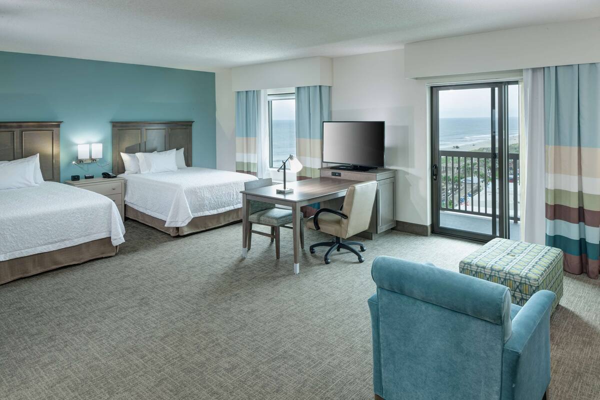 A hotel room with two beds, a desk with a TV, a chair, and a view of the ocean through the balcony.