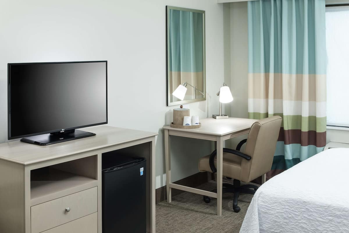 A hotel room with a desk, chair, bed, TV, and a trash bin.