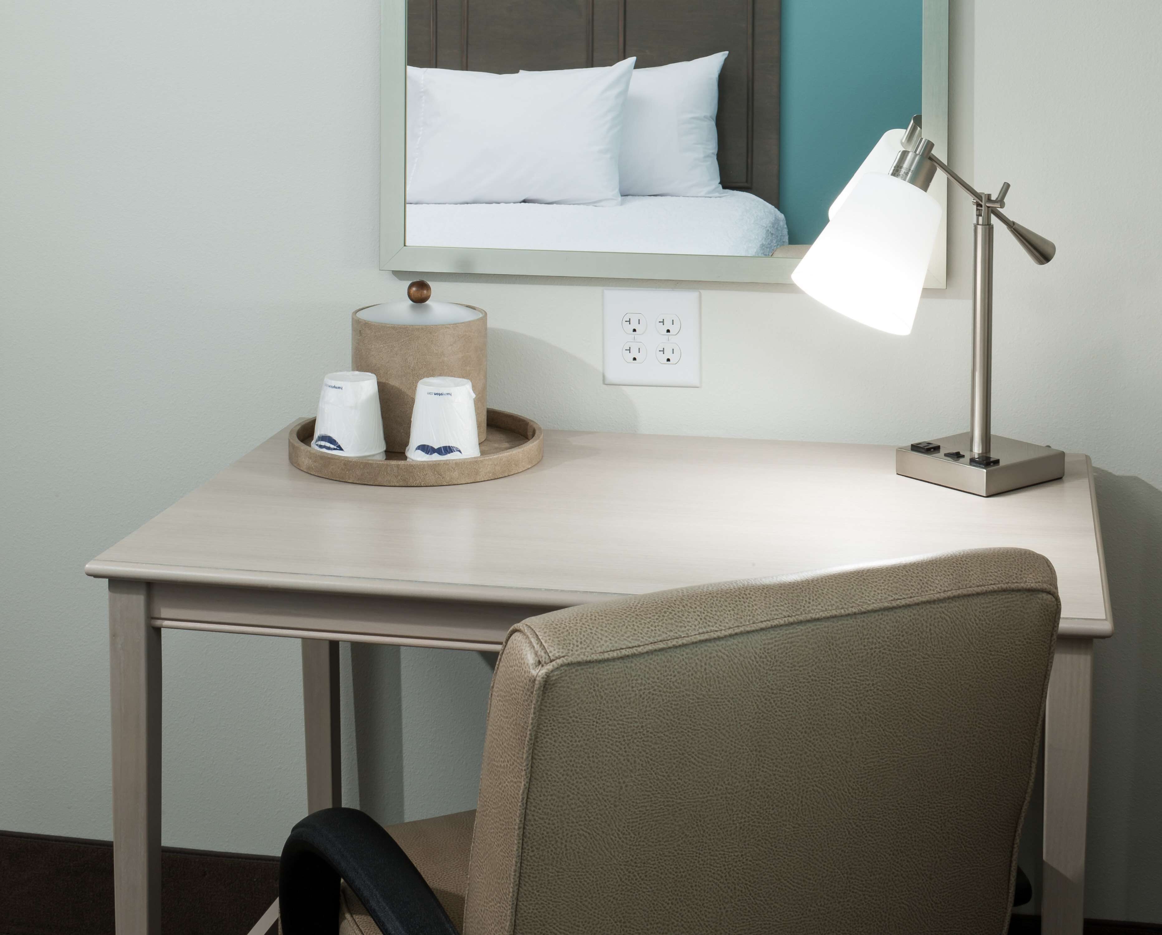 Una scrivania in camera d'albergo con una lampada, uno specchio, un vassoio con tazze e un letto con lenzuola bianche.