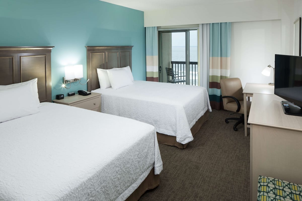 A hotel room with two beds, a desk with a TV, a chair, and a view of the ocean through a balcony.