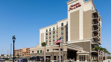 A Hampton Inn & Suites hotel with a modern design, featuring a large American flag, a welcoming entrance, and a parking area with several cars.