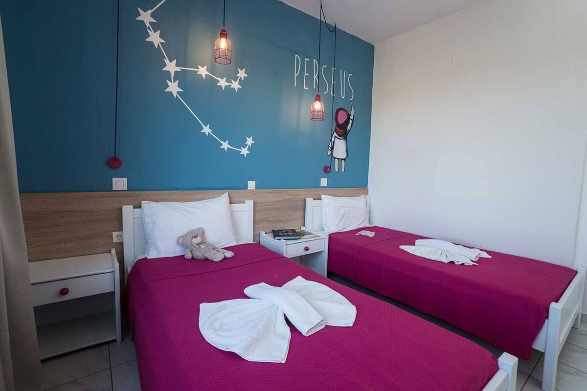Hotellihuone, jossa on kaksi sänkyä. Kummassakin sängyssä on vaaleanpunainen peitto ja valkoiset pyyhkeet. Seinällä on suuri seinämaalaus, jossa on sana ”PERSEUS” ja hahmo, joka pitää palloa.