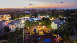 A playground with slides and climbing structures, surrounded by residential buildings and a view of the sea at dusk. - Paradisio Baby and Kinder Hotel (Chania)