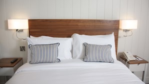 Una cama bien tendida con sábanas blancas y dos almohadas a rayas, flanqueada por mesitas de noche de madera con lámparas y un teléfono.