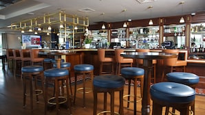 Zona de bar con taburetes de madera, una barra y una variedad de botellas y vasos en estantes.