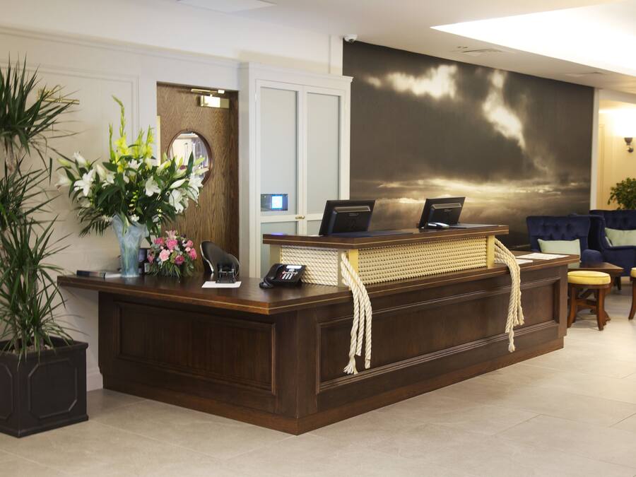 A reception desk with a computer, phone, and a vase of flowers. There is a large painting on the wall and a potted plant to the left.