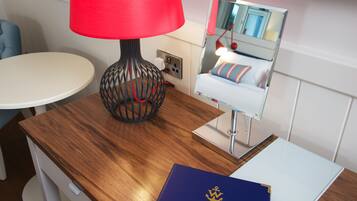 A hotel room with a wooden desk, a red lamp, a mirror reflecting a bed, and a blue booklet on the desk.
