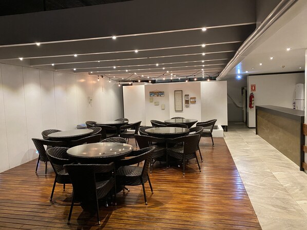A modern indoor dining area with round tables and wicker chairs, wooden flooring, and a ceiling with linear lighting fixtures.