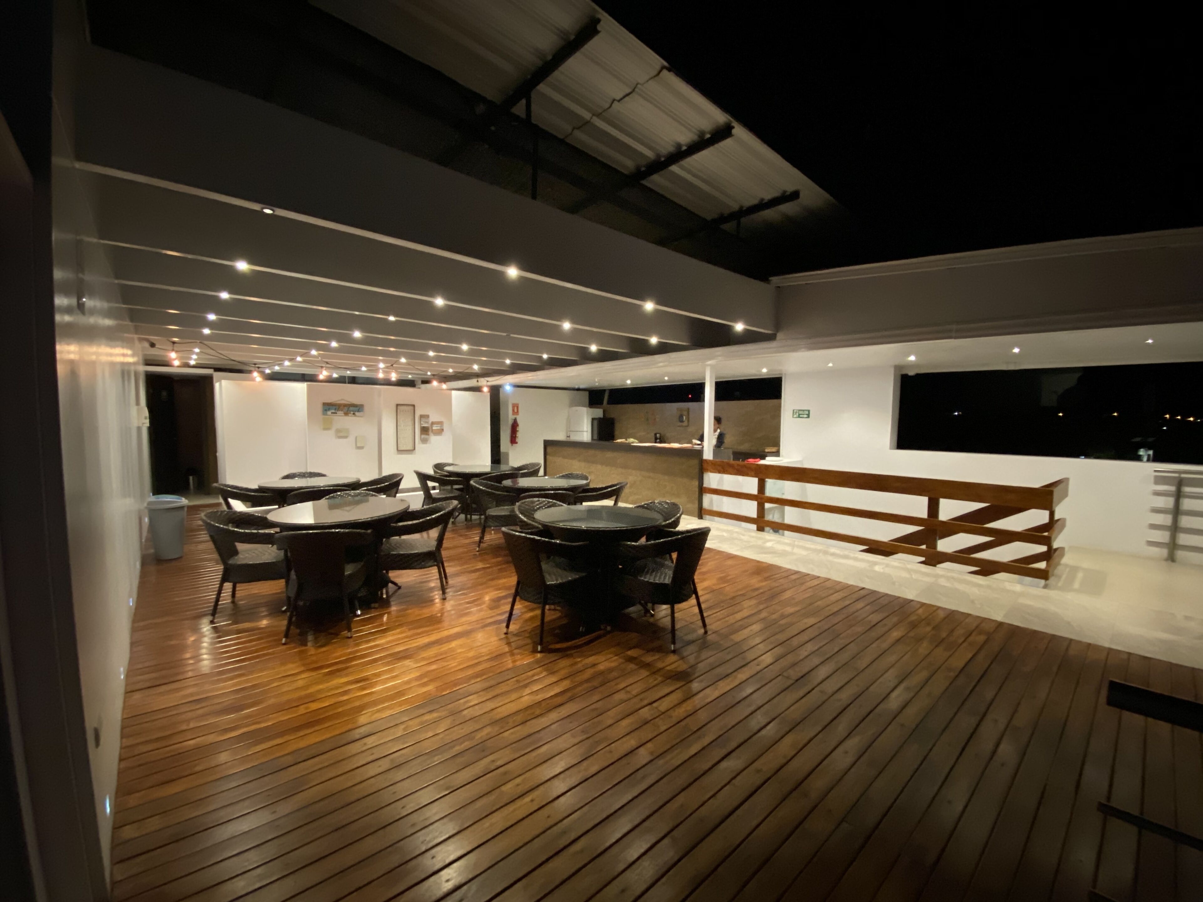 An outdoor dining area with wooden flooring and tables, surrounded by white walls and a wooden fence.