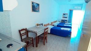 A small hotel room with a dining table, two beds, a painting on the wall, and a window with curtains. - Christi Apartments (Apokoronas)