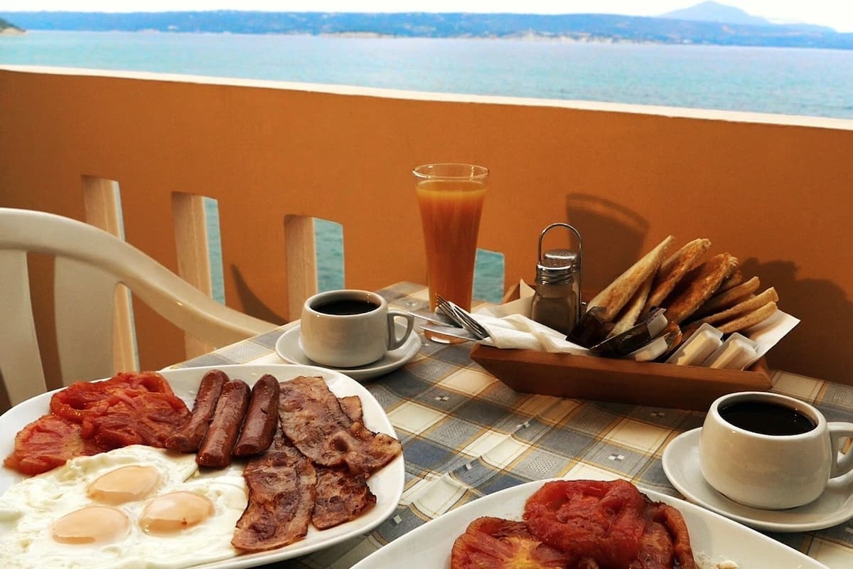 A breakfast table with plates of eggs, bacon, and sausages, a basket of bread, and two cups of coffee.
