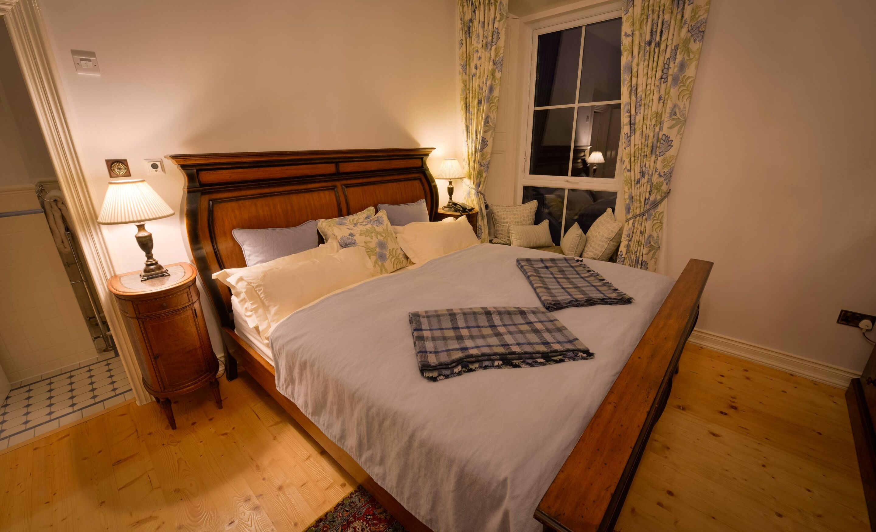 A neatly made bed with a wooden headboard, two bedside lamps, and floral curtains.