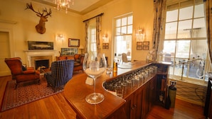 Un coin bar avec un comptoir en bois, des verres et un verre à vin. On y trouve une cheminée surmontée d’un tableau encadré, et une tête de cerf fixée au mur.