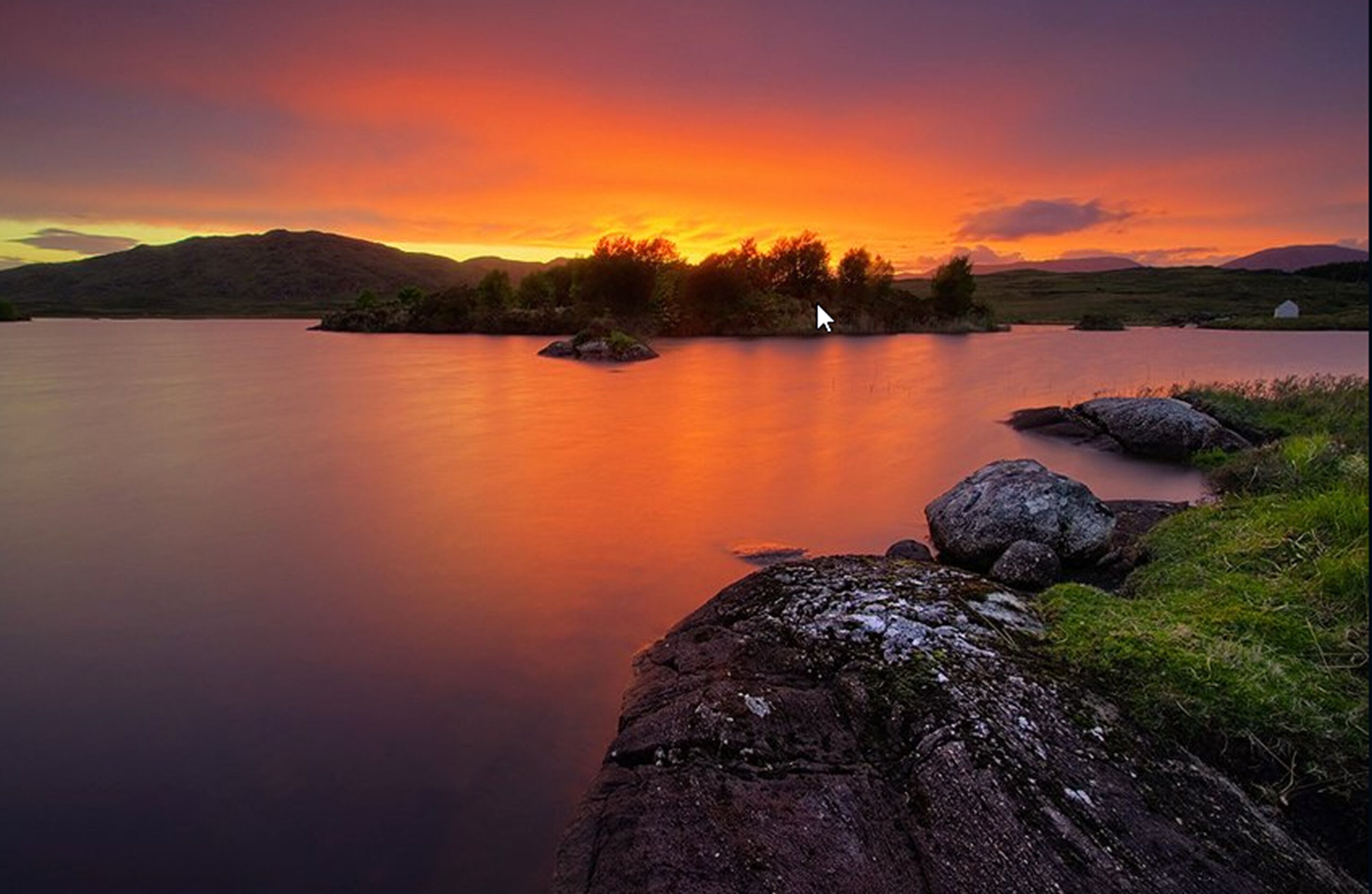 A calm lake with a sunset, surrounded by hills and a small island.