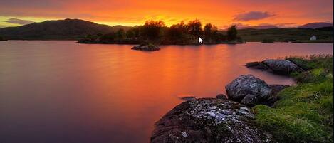 A calm lake with a sunset, surrounded by hills and a small island.