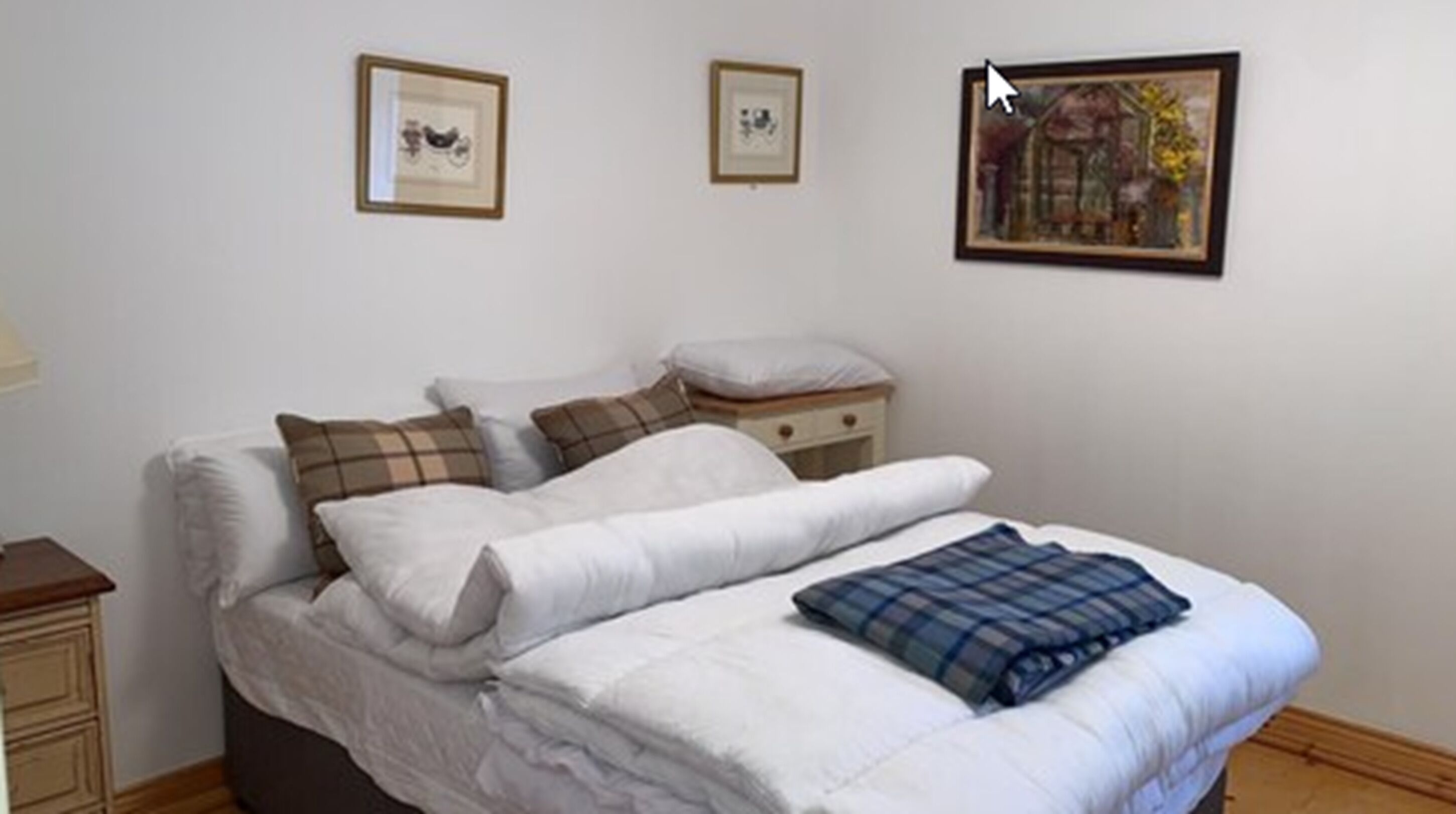 A neatly made bed with white and plaid bedding, a bedside table with a lamp, and two framed pictures on the wall.