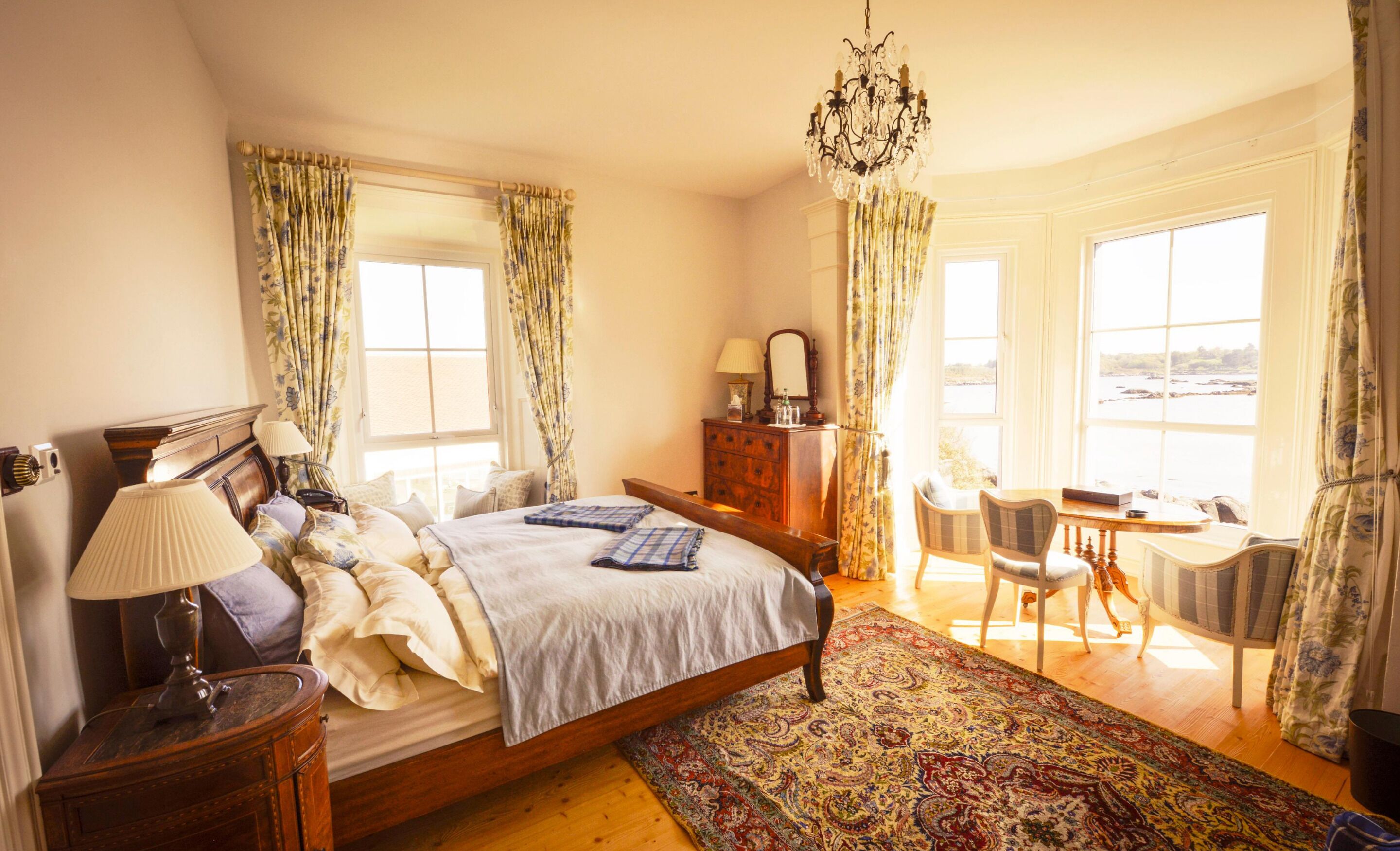 Ein Schlafzimmer mit einem großen Bett, einem Schminktisch, einem Schreibtisch mit Stühlen, einer Chandelüster und großen Fenstern.
