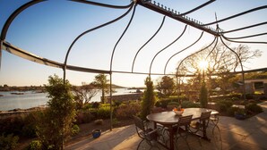 Une terrasse extérieure avec une table ronde et des chaises, entourée de verdure et offrant une vue sur une étendue d’eau.