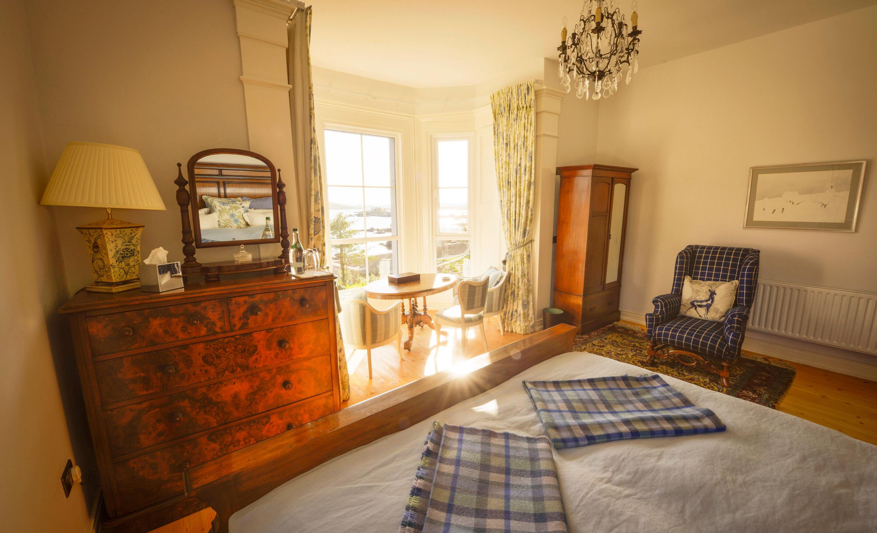 Une chambre avec une commode en bois, un lit recouvert d’une couverture à carreaux, une chaise, une table et une vue sur l’extérieur par une fenêtre.